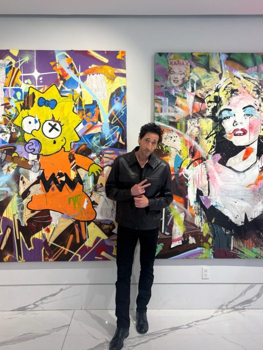 Adrien Brody standing in front of his artworks featuring Marilyn and Maggie Simpson 